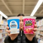 Shopper comparing plain and flavored yogurt labels in store, wondering is flavored yogurt healthy while checking sugar and protein.